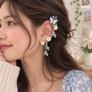 May include: A crochet ear cuff featuring a blue butterfly at the top, followed by a vine of white and blue flowers with small white beads. The intricate thread work follows the curve of the ear, showcasing delicate floral details in blue, white, and green.