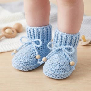 Può includere: Un paio di scarpine per neonati color azzurro chiaro realizzate in filato lavorato a maglia. Ogni scarpina presenta un bordo a coste, un laccetto decorativo con fiocco sul davanti e piccole perline di legno alle estremità. Le scarpine sono indossate.
