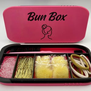 May include: A rectangular pink storage case with the text Bun Box and a line art icon of a hair bun on the lid. The open base is black with four compartments containing clear hair elastics, gold bobby pins, yellow hair sponges, and beige hair ties.