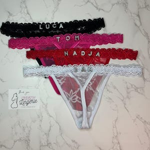 Op de afbeelding: Vier strings in zwart, roze, rood en wit, elk voorzien van een kanten tailleband met strass-letters die LUCA, TOM, NADJA en DAN spellen. Een kleine witte kaart met de tekst Thank you COZY Lingerie is in de buurt geplaatst.