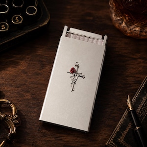 May include: A rectangular silver metal cigarette case featuring a centered embossed design of a cross entwined with thorny vines and a single small red rose. The case is partially open at the top, revealing a row of white cigarettes inside.