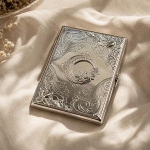 May include: A rectangular silver-toned metal cigarette case featuring an intricate embossed scrollwork pattern across the surface. The center displays a crescent moon and star motif embellished with small clear crystals and white faux pearls.