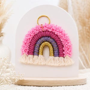Puede incluir: Un colgante de pared de arcoíris de macramé con un anillo de metal dorado en la parte superior. El arco consta de capas de hilo rosa texturizado, cordón morado oscuro y un arco central envuelto en hilo dorado metálico, con flecos color crema.