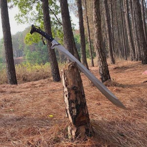 Pode incluir: Uma espada longa e curva com uma lâmina de metal prateado e um cabo preto com um pomo distinto. A espada está posicionada na diagonal, com a ponta apoiada no topo de um toco de árvore vertical e rugoso em uma floresta.