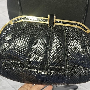 May include: A black clutch bag with a glossy, textured snakeskin pattern. The bag features a curved, gold-toned metal frame along the top edge and a central gold-toned clasp closure. The material has a reflective finish and a structured shape.