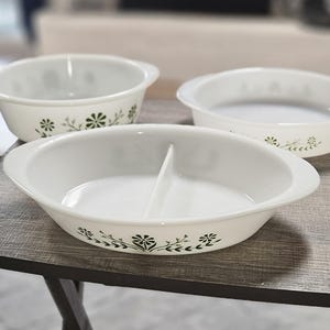 Könnte beinhalten: Drei weiße Glasbackformen mit integrierten Griffen und einem grünen Blumenmuster. Eine Form ist oval mit einer mittigen Unterteilung, während die anderen beiden rund sind. Das grüne Design zeigt stilisierte Blumen und Blätter an der Außenseite.
