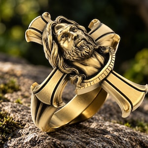 Könnte beinhalten: Ein goldfarbener Ring mit einer dreidimensionalen, skulptierten Büste eines bärtigen Mannes mit langem Haar, die auf einer kreuzförmigen Fassung zentriert ist. Die Metalloberfläche hat ein gebürstetes Finish mit polierten Akzenten.