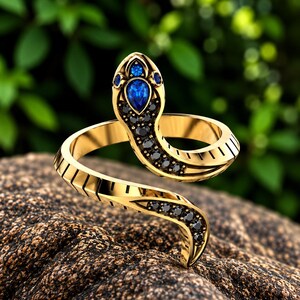May include: A gold snake-shaped ring featuring a pear-cut blue gemstone on the head, accented by small black stones along the head and the winding body. The band has a textured, segmented design that mimics the appearance of snake scales.
