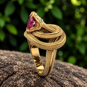 May include: A gold ring shaped like a coiled snake with a textured, beaded surface. The snake head features a large, marquise-cut deep red gemstone in its mouth and a small, clear round accent stone set near the eye area on the side of the head.