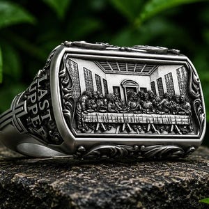 Könnte beinhalten: Ein silberfarbener Ring mit einer rechteckigen Vorderseite, die ein detailliertes Relief des Abendmahls zeigt. Die Seite des Rings ist mit einem eingravierten Kreuz und dem Text LAST SUPPER in Großbuchstaben sowie Verzierungen versehen.
