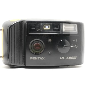 Peut inclure: Un appareil photo argentique compact Pentax PC-606W noir doté d'un objectif frontal, d'un flash intégré, d'une fenêtre de mise au point automatique et d'un interrupteur circulaire. Le boîtier présente une zone de préhension texturée et une dragonne.