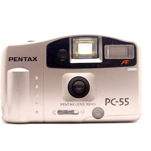 Peut inclure: Un appareil photo argentique 35 mm Pentax PC-55 avec un objectif Pentax 30 mm, un système autofocus, un flash intégré et un viseur. La face avant du boîtier affiche la marque Pentax, le modèle PC-55 et un logo AF.