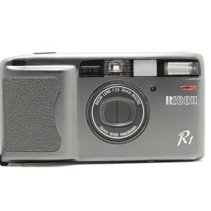 May include: A dark gray Ricoh R1 compact film camera featuring a Ricoh Lens 1:3.5 30mm Macro and 24mm Wide Panorama lens. The front body includes a textured black grip on the left side, a flash unit, a viewfinder window, and the Ricoh logo.