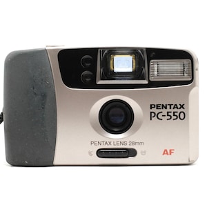 May include: A silver and black Pentax PC-550 point-and-shoot camera featuring a Pentax Lens 28mm, an autofocus system, a built-in flash, and a viewfinder. The camera body has a textured black grip on the left side and a black wrist strap attached.