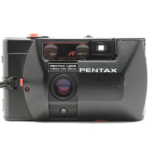 Peut inclure: Un appareil photo argentique 35mm noir Pentax doté d'un objectif fixe 35mm f/2.8, d'un système autofocus et d'un déclencheur rouge. L'avant affiche le texte PENTAX LENS f=35mm 1:2.8 0.7m-infinity, AF AUTO FOCUS et la marque PENTAX.