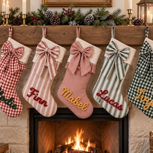 May include: Five personalised Christmas stockings hanging in a row. From left to right: a red and white gingham stocking with 'Elodie', a red and white striped stocking with 'Finn', a solid pink stocking with 'Mabel', a green and white striped stocking with 'Lucas', and a green and white gingham stocking with 'Ivy'. Each features a matching fabric bow at the top.