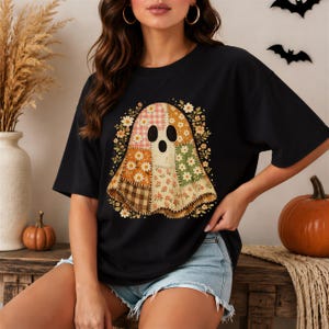 May include: A black short-sleeve t-shirt featuring a central graphic of a ghost composed of various patchwork fabric patterns, including gingham, floral, and plaid prints in shades of orange, brown, green, and cream, surrounded by small flowers.