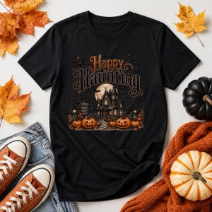 May include: A black t-shirt featuring a graphic print of a haunted house under a full moon with the text Happy Haunting. The design includes orange pumpkins, bare trees, and flying bats, creating a detailed Halloween-themed illustration.