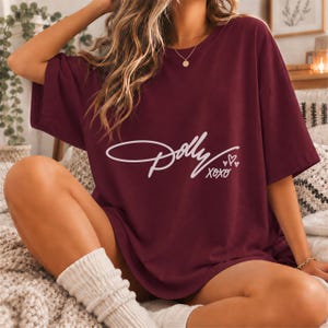 May include: A maroon short-sleeved t-shirt featuring the white cursive text "Dolly" followed by "xoxo" and three small heart icons printed on the center of the chest. The garment has a crew neckline and a loose, relaxed fit throughout the body.