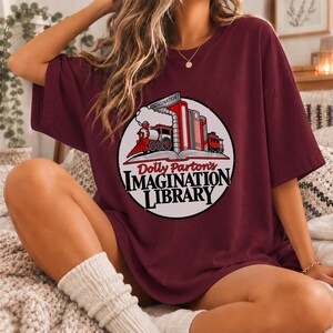 Puede incluir: Una camiseta de manga corta color granate con un gráfico circular en el pecho. El diseño muestra un tren de vapor saliendo de un libro abierto junto a una fila de libros, con el texto Dolly Parton's IMAGINATION LIBRARY impreso debajo.