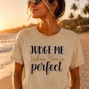 Könnte beinhalten: Ein beiges Kurzarm-T-Shirt mit dem schwarzen und goldenen Text "JUDGE ME When You're perfect" auf der Vorderseite. Das T-Shirt hat einen Rundhalsausschnitt und wird mit einer blauen Jeans kombiniert, was einen lässigen Stil mit hochgekrempelten Ärmeln zeigt.