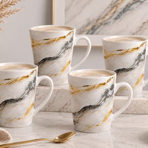 May include: Four ceramic mugs featuring a white base with swirling gray and gold metallic accents. Each mug has a handle on the side and is filled with a light brown beverage. The mugs are arranged in a group on a white marble surface.