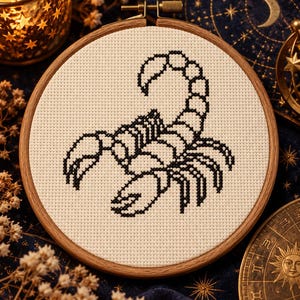 以下が含まれることがあります： オフホワイトのアイーダ布が入った円形の木製刺繍枠。布の中央には、黒い糸でステッチされたサソリのクロスステッチデザインがあり、湾曲した尾、2つの大きなハサミ、8本の脚が描かれています。