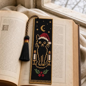 Op de afbeelding: Een rechthoekige kruissteek boekenlegger op een zwarte achtergrond met een zwarte kat die een rood-witte kerstmuts draagt, geflankeerd door twee witte kaarsen, een gele maansikkel, sterren en rode hulstbessen met groene bladeren aan de onderkant.