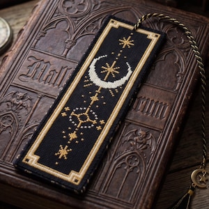 Puede incluir: Un marcapáginas rectangular negro con motivos celestiales en punto de cruz dorados y blancos, incluyendo una luna creciente y estrellas. El marcapáginas tiene un borde geométrico dorado y un cordón trenzado negro y dorado con un dije de luna.