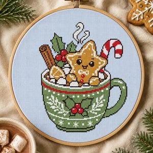 May include: A cross-stitch embroidery in a wooden hoop featuring a green mug with a holly pattern filled with hot cocoa, marshmallows, a cinnamon stick, a candy cane, and a smiling star-shaped gingerbread cookie on a light blue background.