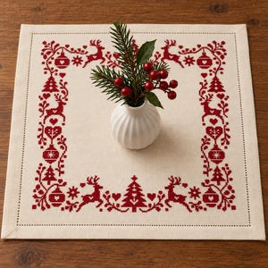 May include: A square off-white fabric mat featuring a red cross-stitch border pattern of reindeer, Christmas trees, hearts, and ornaments. A small white ribbed ceramic vase sits in the center, holding a sprig of pine needles and red berries.