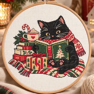 May include: A circular wooden embroidery hoop containing a cross-stitch design of a black cat wearing a red and white patterned scarf. The cat is reading a green book with a Christmas tree illustration, sitting beside a stack of books, a gift, and a mug.