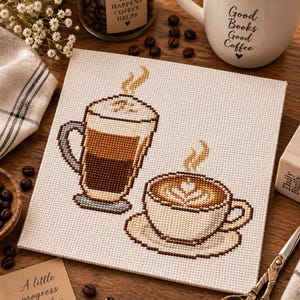 Könnte beinhalten: Eine quadratische Kreuzstichstickerei auf weißem Stoff, die ein hohes geschichtetes Kaffeegetränk und eine breite Kaffeetasse mit Latte-Art-Muster zeigt. Aus beiden Getränken steigt Dampf auf. Das Design verwendet braune, beige und graue Garne.