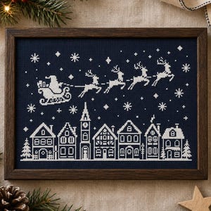 Pode incluir: Um bordado em ponto cruz emoldurado com um design de fio branco sobre tecido azul-marinho escuro. O padrão retrata uma cena de aldeia de inverno com casas e uma torre de relógio sob a silhueta do Pai Natal num trenó puxado por renas.