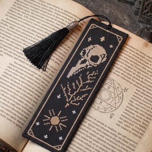 Puede incluir: Un marcapáginas rectangular de tela negra con un diseño de punto de cruz en hilo beige y blanco. El patrón incluye un cráneo de ave, una rama con hojas, un sol y pequeñas estrellas. Una borla negra con un casquillo plateado está unida.