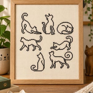 May include: A framed cross-stitch embroidery on cream-colored Aida cloth featuring seven distinct black-thread outlines of cats in various poses, including stretching, sitting, and curled up, all enclosed within a light-toned wooden frame.