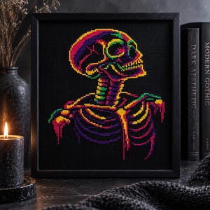 May include: A square black frame containing a cross-stitch embroidery of a human skeleton. The skeleton is rendered in vibrant neon colors including yellow, green, purple, and pink against a solid black fabric background, creating a high-contrast effect.