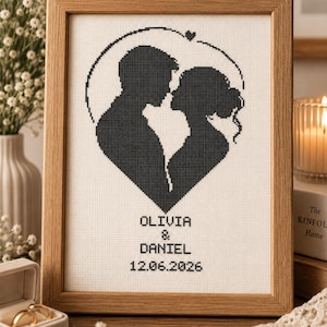 May include: A framed cross-stitch artwork featuring a black silhouette of a couple facing each other within a heart-shaped outline. Below the figures, the text reads OLIVIA & DANIEL and the date 12.06.2026, all stitched in black on white fabric.