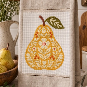 May include: A beige linen kitchen towel featuring a centred cross-stitch design of a yellow pear. The pear is decorated with white floral patterns, small orange accents, and a single green leaf. The towel has a hemmed border with decorative stitching.