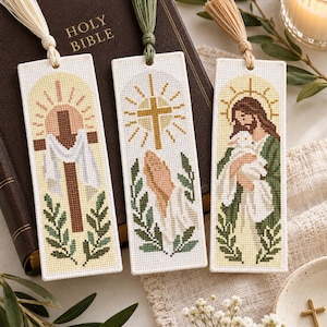 May include: Three rectangular cross-stitch bookmarks featuring religious imagery. The first shows a cross with a white cloth, the second shows praying hands with a golden cross, and the third shows a figure holding a lamb. Each has a colored tassel.