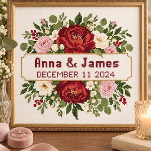 May include: A square framed cross-stitch embroidery featuring a floral wreath of red, pink, and white flowers surrounding a central rectangular banner. The text reads Anna & James and DECEMBER 11 2024 in dark red and black stitched lettering.