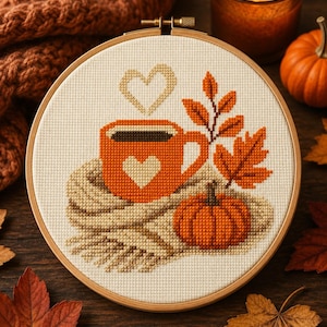 May include: A cross-stitch embroidery in a circular wooden hoop featuring an orange mug with a heart, a pumpkin, and autumn leaves on a beige fabric background. Steam in the shape of a heart rises from the mug, which rests on a folded scarf.