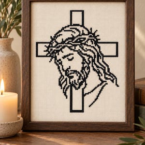 May include: A framed cross-stitch artwork featuring a black thread design of a person wearing a crown of thorns in front of a cross, set against a cream-colored fabric background within a dark brown wooden frame.