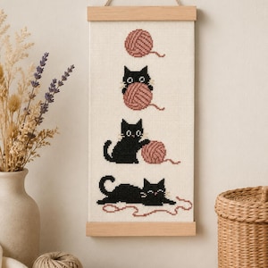 Peut inclure: Une tenture murale en point de croix représentant un chat noir interagissant avec une pelote de laine rose poudré en trois étapes. La broderie est montée sur un cadre en bois clair avec une cordelette, sur un fond en tissu blanc.