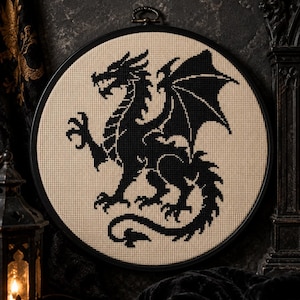 Könnte beinhalten: Ein runder Stickrahmen mit einem Kreuzstichmotiv, das die Silhouette eines schwarzen Drachen auf beigem Stoff zeigt. Der Drache ist im Profil mit ausgebreiteten Flügeln, scharfen Krallen und einem langen, geschwungenen Schwanz dargestellt.