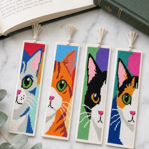 Puede incluir: Cuatro marcapáginas rectangulares de punto de cruz con retratos de gatos. Cada marcapáginas muestra una cara felina diferente en colores vibrantes sobre fondos geométricos, rematados con una pequeña borla color crema en el borde superior.