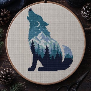 May include: A circular wooden embroidery hoop containing a cross-stitch pattern of a howling wolf. The wolf silhouette is filled with a landscape featuring dark blue pine trees, light blue snow-capped mountains, a crescent moon, and white stars.