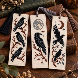 May include: Three rectangular cross-stitch bookmarks on cream-coloured fabric. The left features three black ravens on brown branches with a black tassel. The centre shows a raven, moon, and stars with a grey tassel. The right depicts a flying raven, a crescent moon, and a perched raven with a brown tassel.