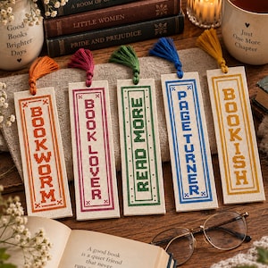 May include: Five rectangular cross-stitch bookmarks on a beige fabric surface. Each bookmark features a colored tassel and embroidered text: orange BOOKWORM, pink BOOK LOVER, green READ MORE, blue PAGE TURNER, and yellow BOOKISH, all in a block font.