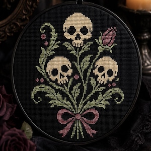 May include: A circular embroidery hoop containing a cross-stitch pattern on black fabric. The design features three beige skulls arranged among green leafy stems and small pink flowers, all tied together at the base with a pink ribbon bow.